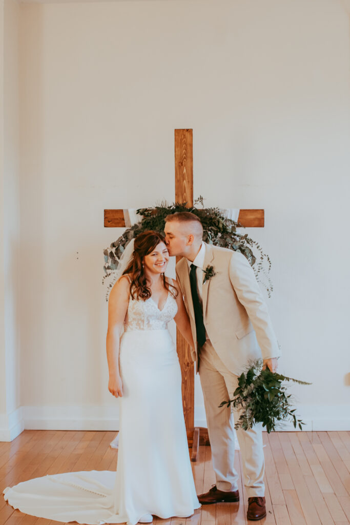 groom kissing bride on the cheek in front of cross