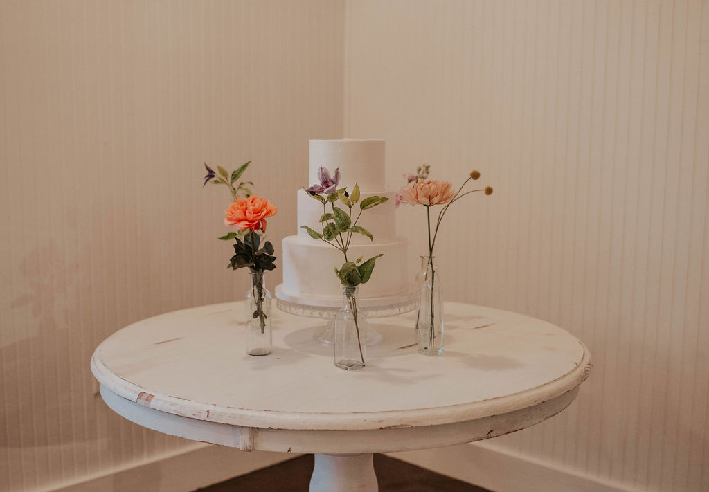 simple wedding cake surrounded by single flowers in small vases